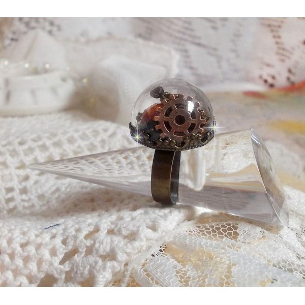 Bague Majest&eacute; Steampunk aux rouages m&eacute;caniques &agrave; laquelle s&rsquo;ajoute une cl&eacute; et un strass de couleur Siam et un strass en verre Dor&eacute;. Le tout est mis sous un globe en verre sur une bague r&eacute;glable laiton