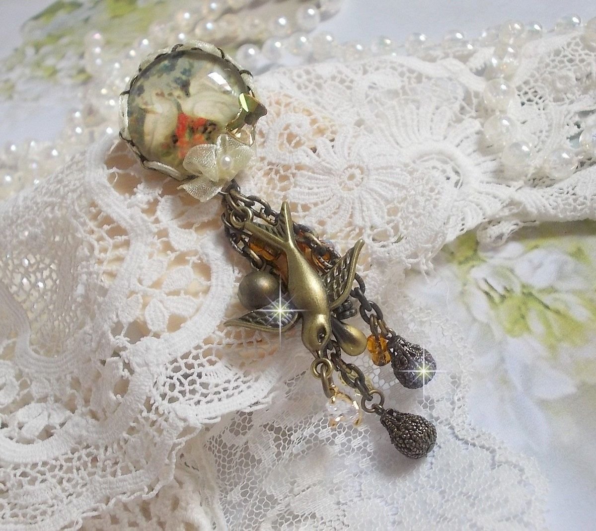 Broche Colombes aux plumages blancs avec des breloques filigranées gouttes et lisses, perles rondes et nœud en cristal, ruban organza crème, pendentif plaqué or avec un zircon sur une broche en métal