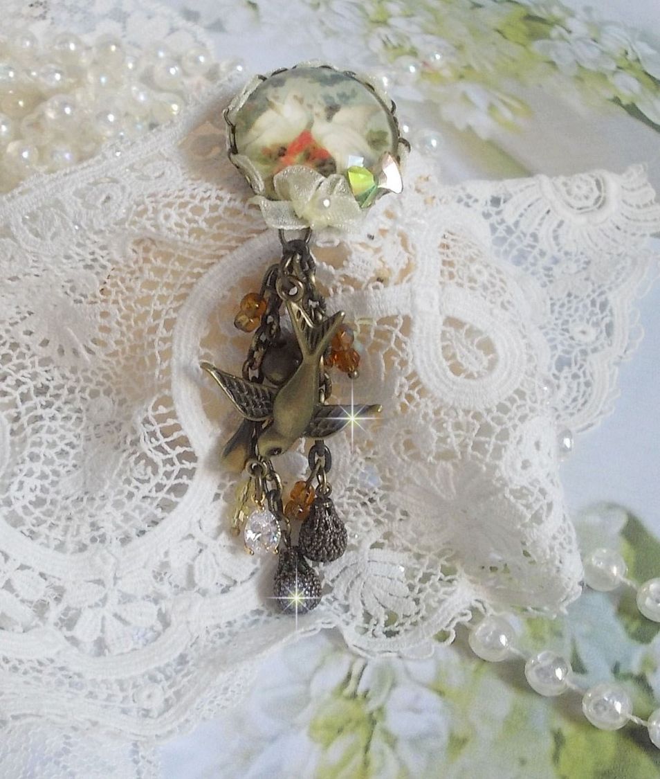 Broche Colombes aux plumages blancs avec des breloques filigranées gouttes et lisses, perles rondes et nœud en cristal, ruban organza crème, pendentif plaqué or avec un zircon sur une broche en métal