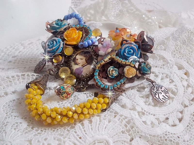 Broche Eclats P&eacute;tillants d&rsquo;inspiration Baroque, orn&eacute;e de fleurs en r&eacute;sine autour d&rsquo;un cabochon strass&eacute;s et perl&eacute;s sur estampes, cristaux, breloques, perle cloisonn&eacute;e, perles jaunes en verre et roses. 