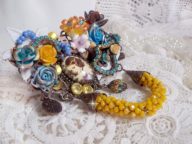 Broche Eclats P&eacute;tillants d&rsquo;inspiration Baroque, orn&eacute;e de fleurs en r&eacute;sine autour d&rsquo;un cabochon strass&eacute;s et perl&eacute;s sur estampes, cristaux, breloques, perle cloisonn&eacute;e, perles jaunes en verre et roses. 