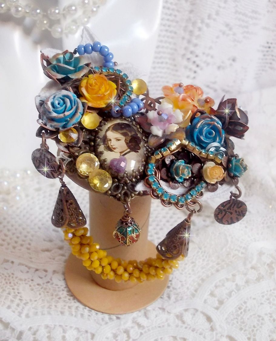 Broche Eclats Pétillants d’inspiration Baroque, ornée de fleurs en résine autour d’un cabochon strassés et perlés sur estampes, cristaux, breloques, perle cloisonnée, perles jaunes en verre et roses. 